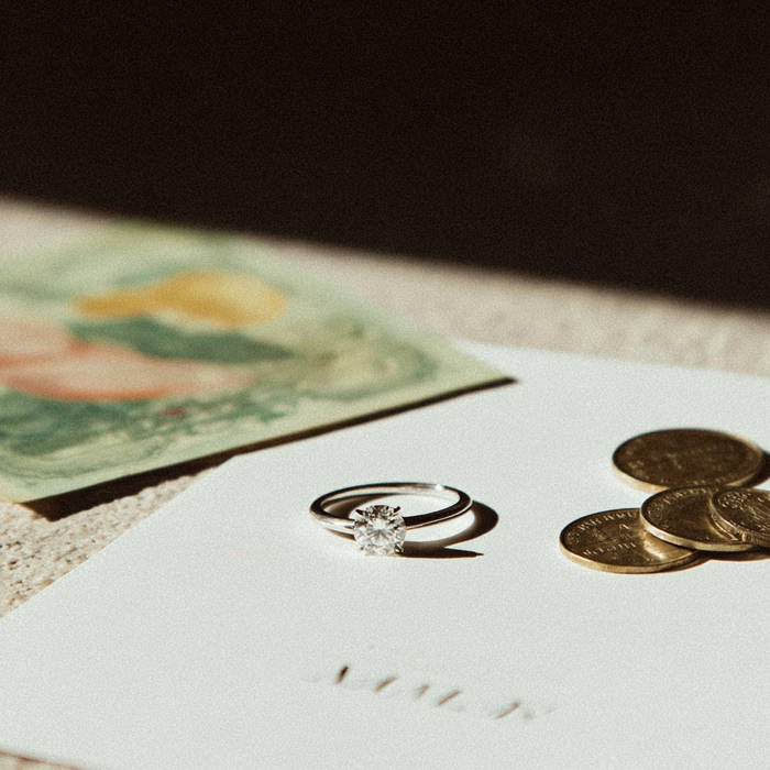 Solitaire diamond engagement ring resting on white paper beside a few coins, with soft sunlight and a blurred illustration in the background