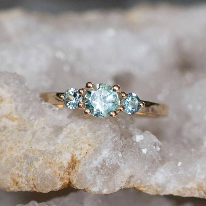 Yellow gold ring with a round pale blue aquamarine center stone flanked by two smaller matching blue gemstones, displayed on a natural crystal surface.