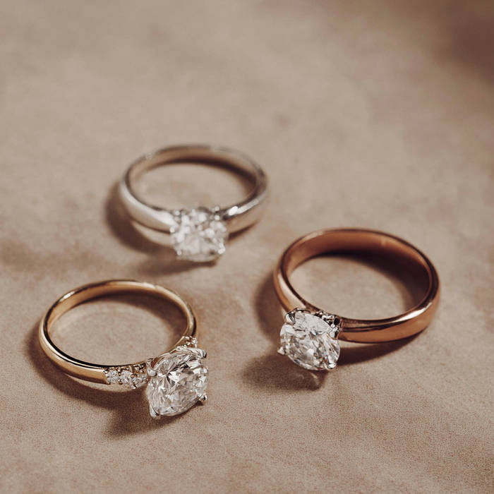 Three engagement rings with solitaire diamonds in yellow, white, and rose gold arranged on a soft beige surface under warm light