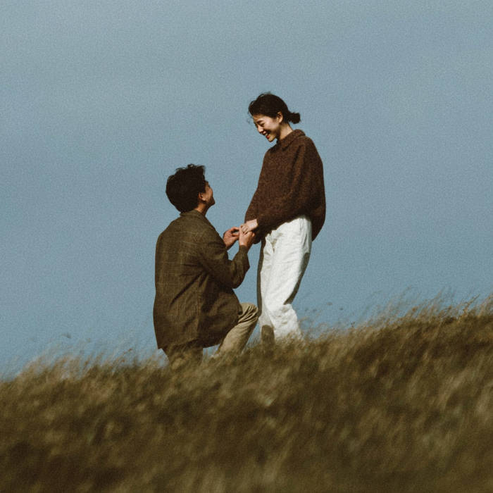 Man kneeling to propose with a bespoke engagement ring on a grassy hill as his partner smiles, blue sky backdrop and natural setting