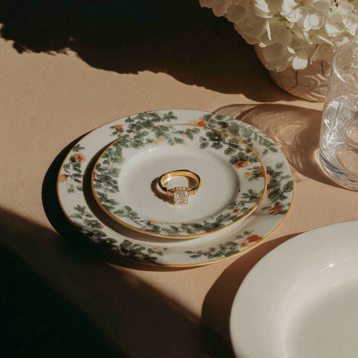 A yellow gold engagement ring with a square-cut diamond sits centered on a small floral-patterned plate, styled on a softly lit table with layered china and glassware creating warm, dramatic shadows.