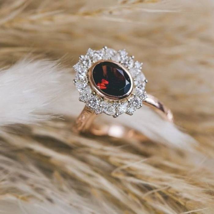 Rose gold ring with an oval deep red garnet center stone surrounded by a halo of small white diamonds, displayed on a soft fur-textured background.