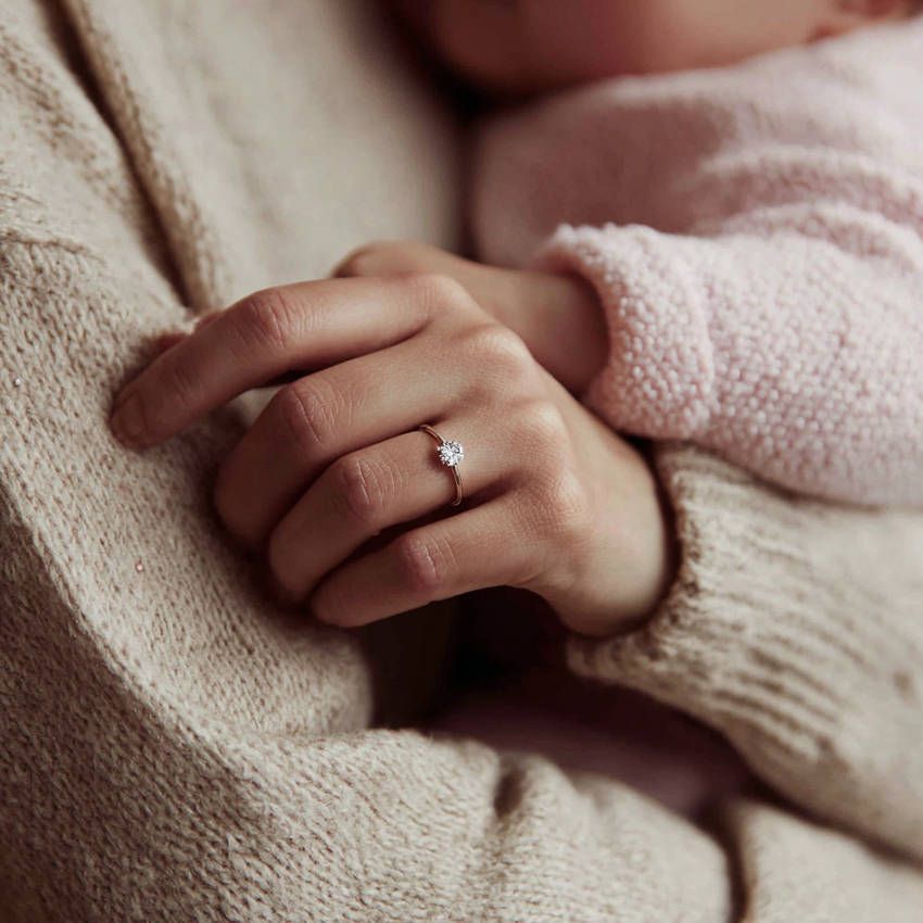 Close-up of a woman holding a baby, wearing a custom solitaire diamond engagement ring on her hand.