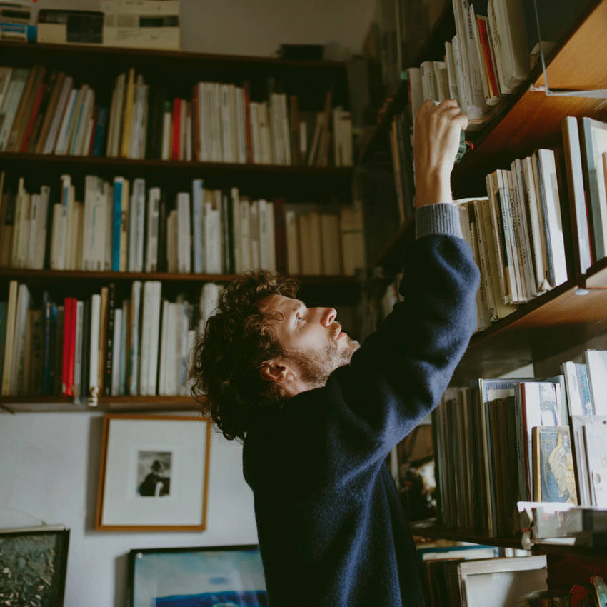 Man reaching for a high bookshelf in a cozy home library, surrounded by books and warm natural light.