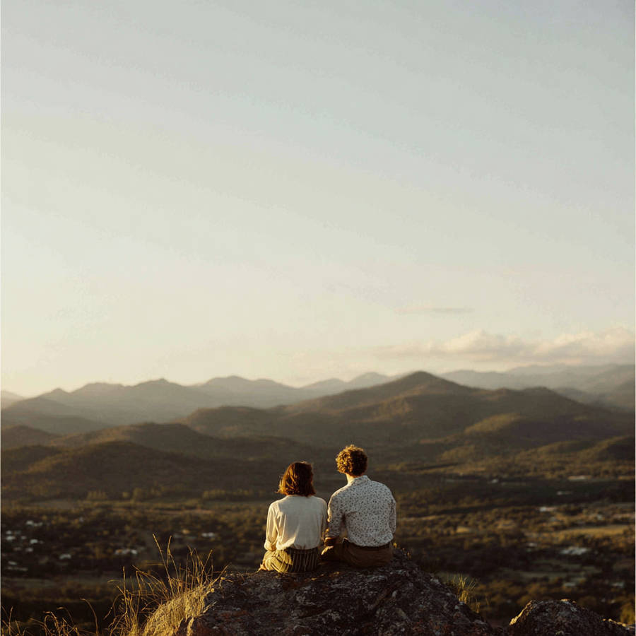 Couple sitting on a rock at sunset, overlooking a wide valley and layered mountains in the distance.
