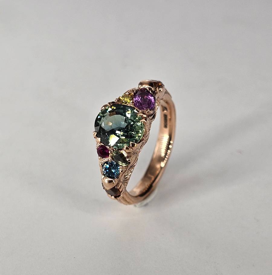 Rose gold ring with a large oval green gemstone surrounded by smaller multicoloured gemstones, including purple, blue, pink, and yellow stones, set in an organic textured band on a light neutral background.