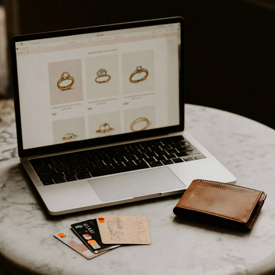Laptop showing engagement ring listings on a jewelry website, with credit cards and a leather wallet on a marble table