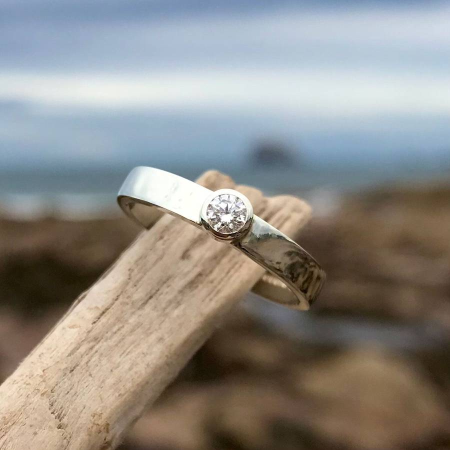 Bespoke bezel-set diamond engagement ring with polished white gold band resting on driftwood by the sea