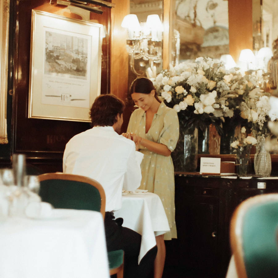 Woman smiling as she places a bespoke engagement ring on her finger during a romantic proposal in an elegant restaurant interior