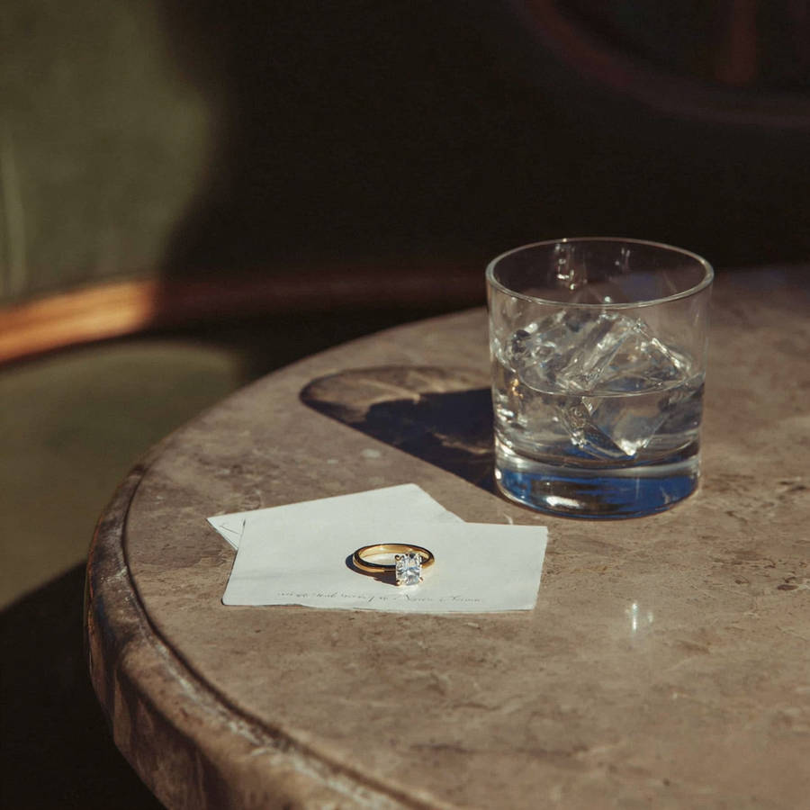 Solitaire diamond engagement ring on a card beside a glass of whiskey on a marble table in warm, moody lighting