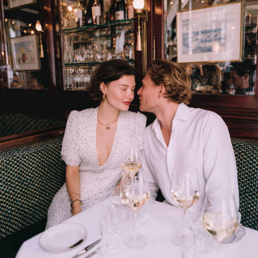 Couple leaning in closely at a cozy bistro table, sharing wine in an intimate restaurant setting with mirrored shelves and warm lighting