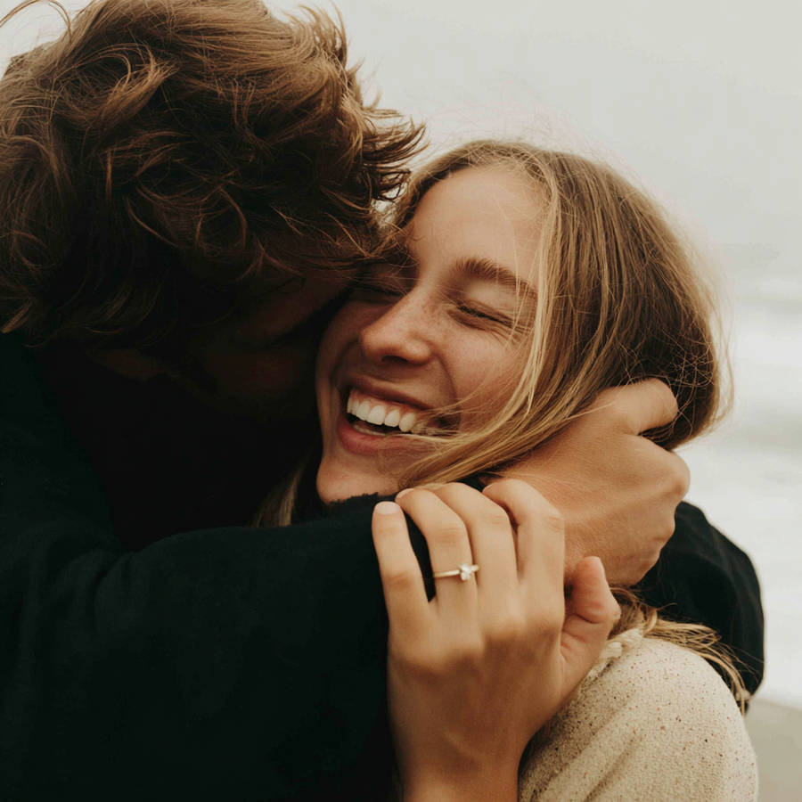 Couple hugging by the sea, smiling as a bespoke engagement ring sparkles on her hand