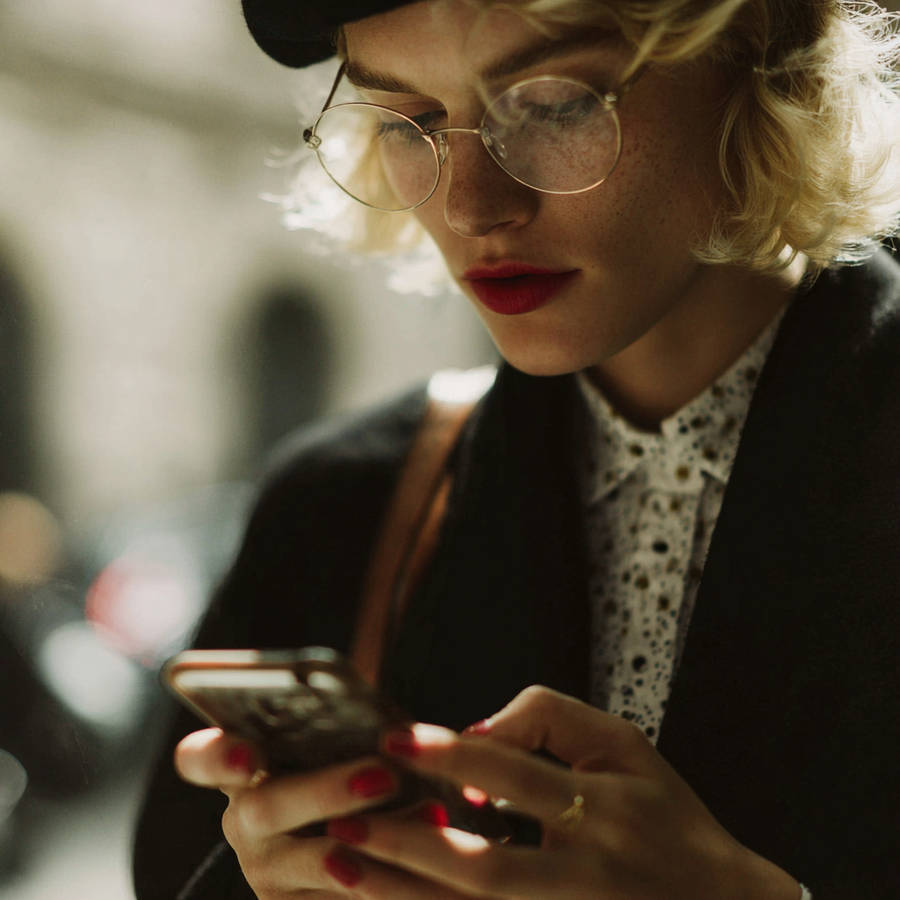 Woman in beret and round glasses typing on a smartphone, red lipstick and nails in soft streetlight.