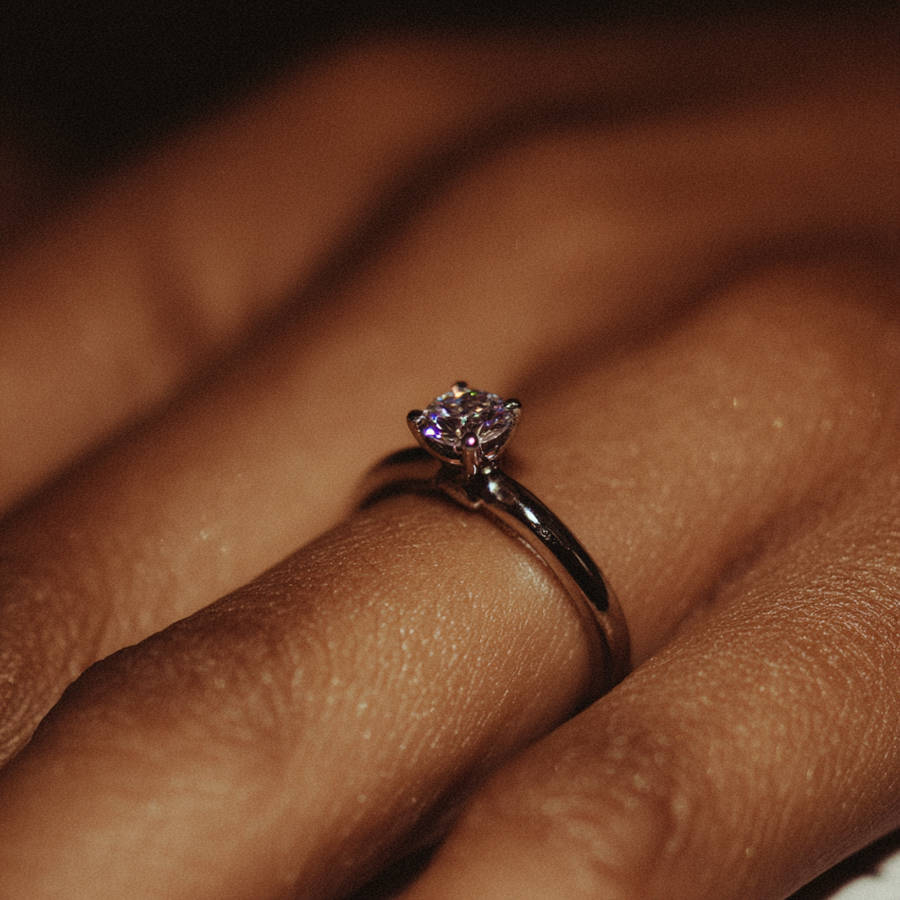 Close-up of a delicate custom solitaire diamond engagement ring on a finger, catching warm light with subtle sparkle
