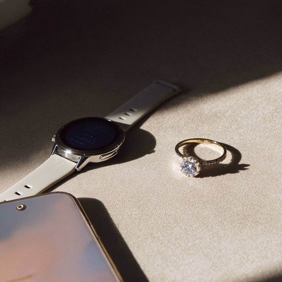 Flat lay of bespoke diamond engagement ring beside smartwatch and phone on soft fabric in warm natural light
