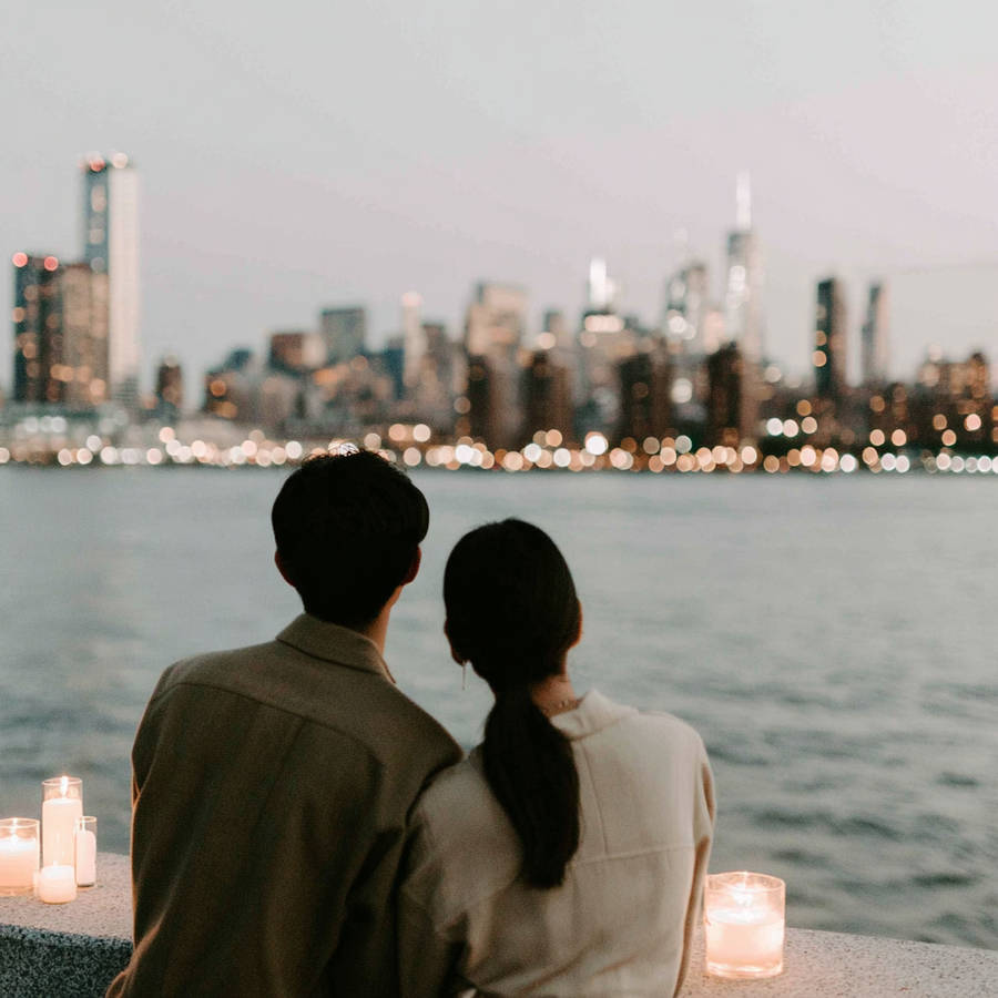 Couple sitting side by side by the water, watching a softly lit city skyline at dusk with candles glowing nearby