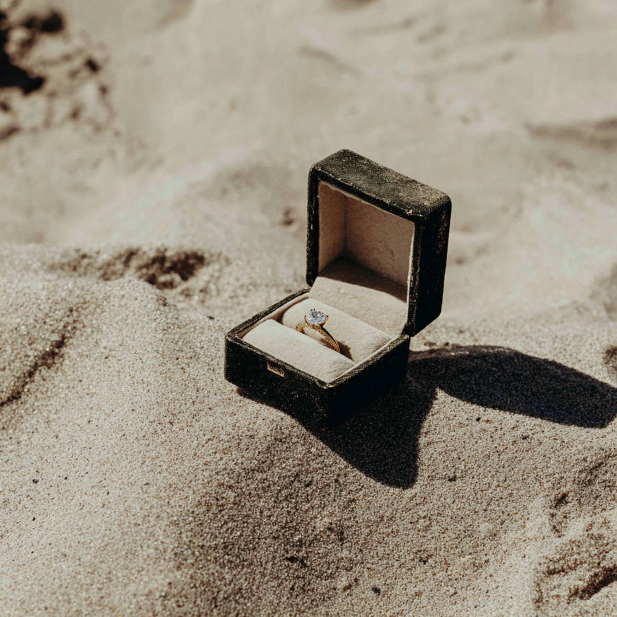 Open ring box holding a bespoke diamond engagement ring on a sandy beach in warm sunlight