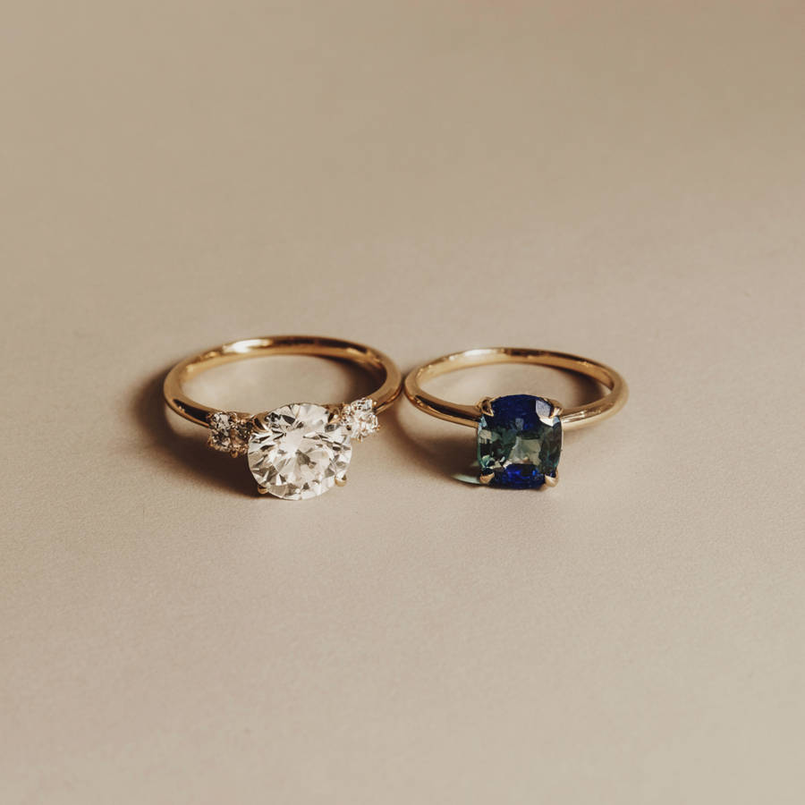 Two gold bespoke rings side by side, one with a round diamond and side stones, the other with a blue cushion-cut gemstone