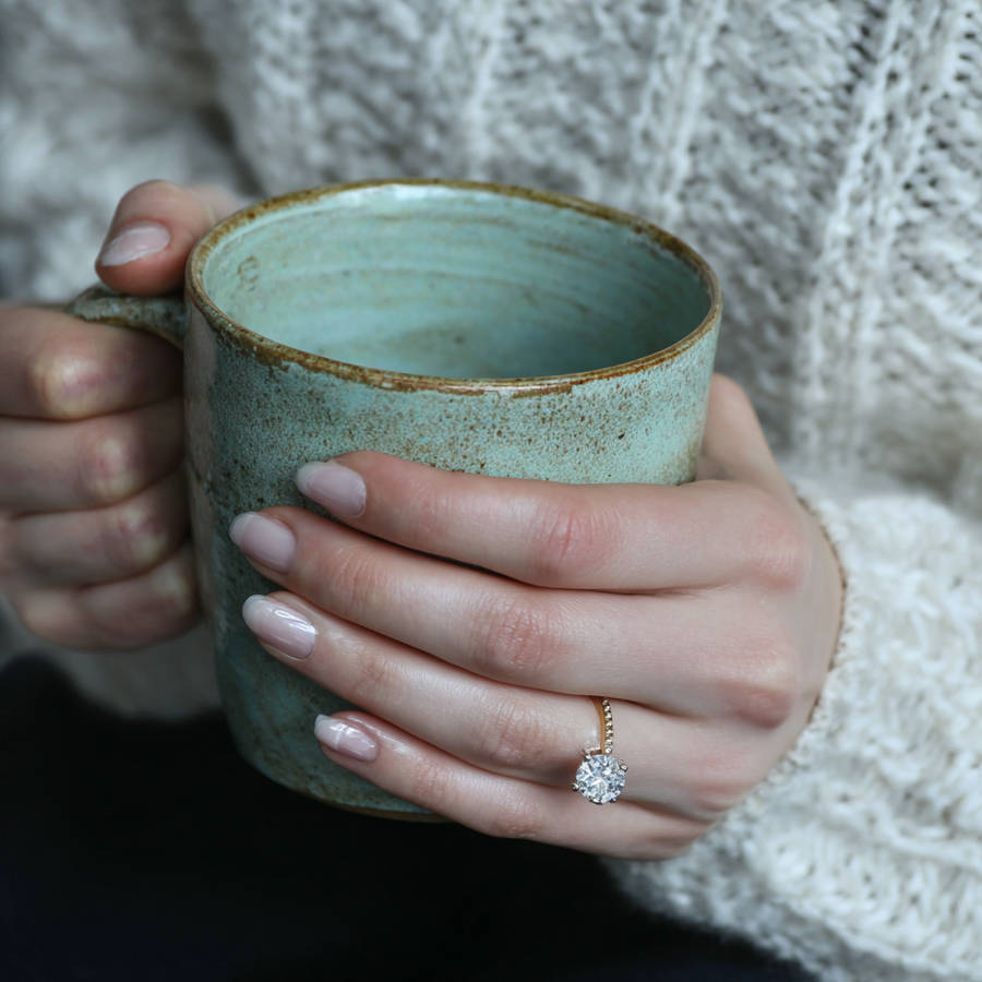 Close-up of a bespoke engagement ring on a hand holding a ceramic mug, with a round diamond on a gold band