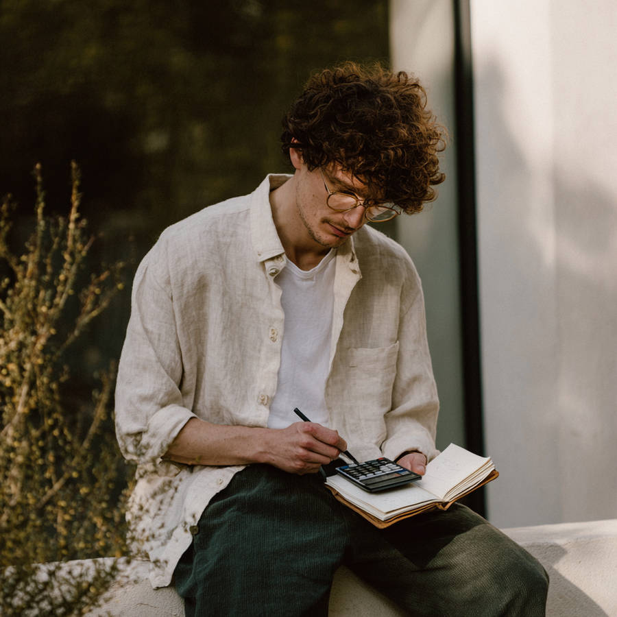 Curly-haired man in glasses uses a calculator and pen over an open notebook, calculating a budget outdoors