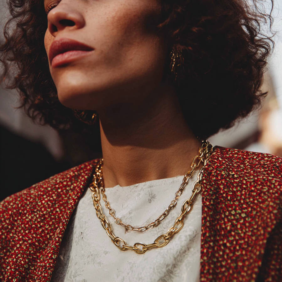 Close-up of a woman wearing layered gold chain necklaces and gold hoop earrings, dressed in a patterned red jacket over a light top, with soft natural light highlighting her face and jewelry.