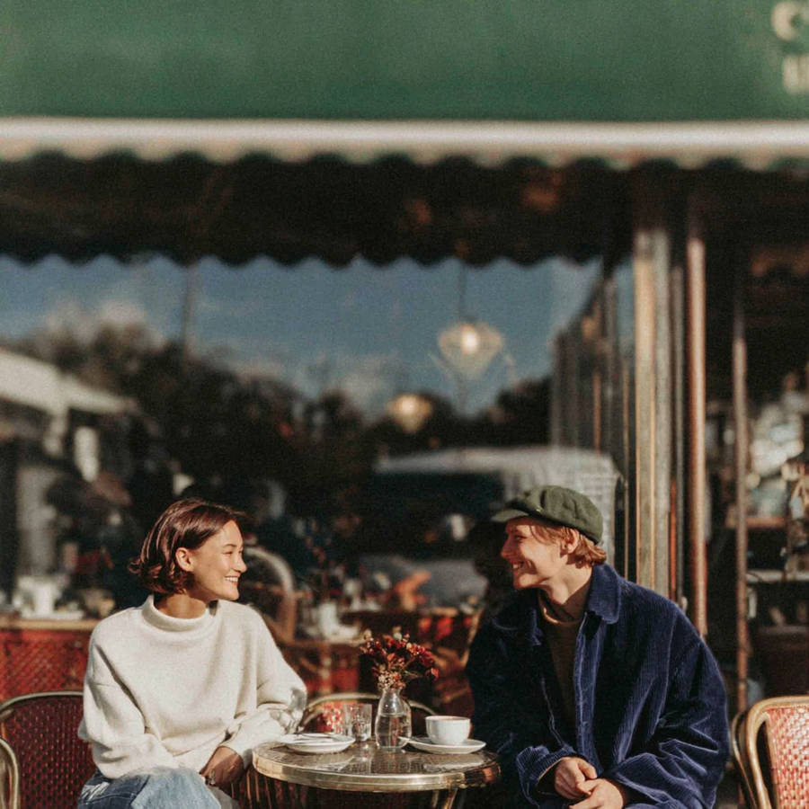 A couple smiles at each other over coffee at an outdoor café table in warm afternoon sunlight