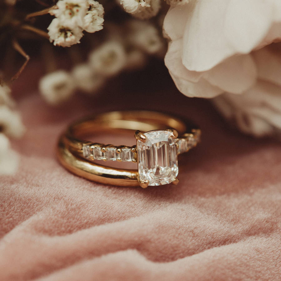 Ultra close-up of a bespoke gold engagement ring with emerald-cut diamond and baguette side stones on soft pink fabric
