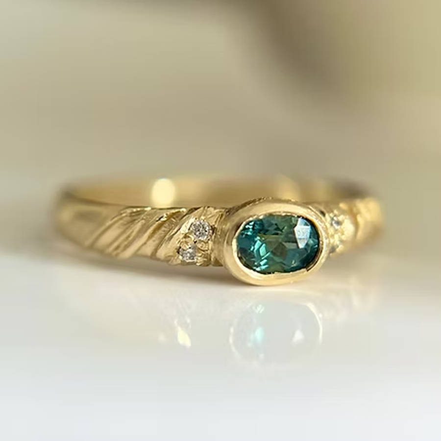 Gold ring with an oval teal gemstone in a smooth bezel setting, accented by small diamonds and subtle engraved details on the band, photographed close up on a light surface
