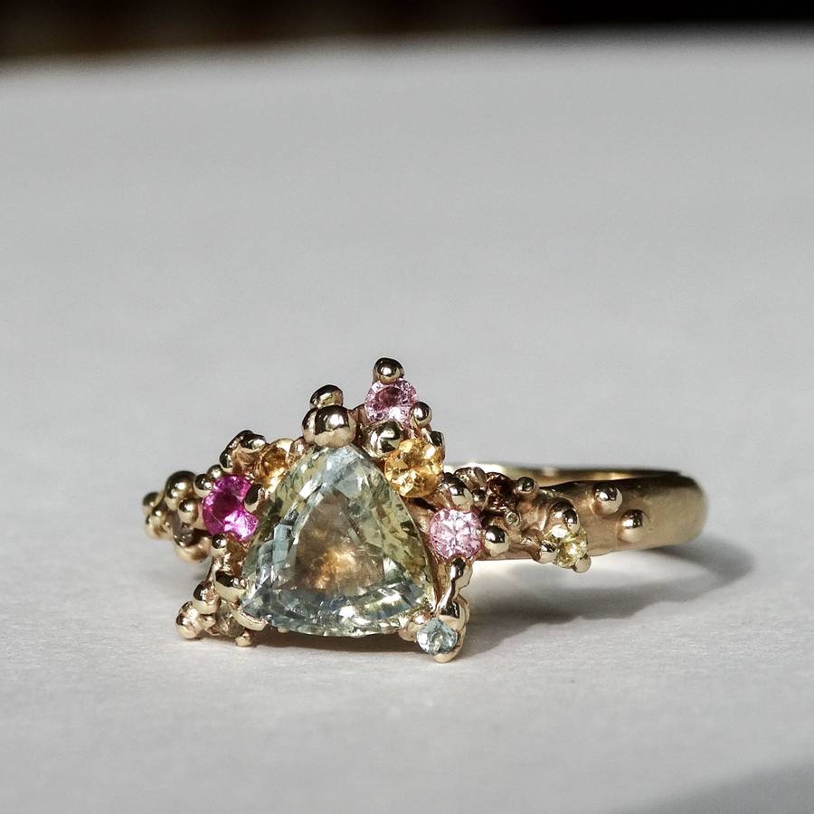 Organic gold ring with a triangular pale green gemstone centre, surrounded by small pink, yellow, and blue gemstones in an irregular cluster setting on a light neutral background.