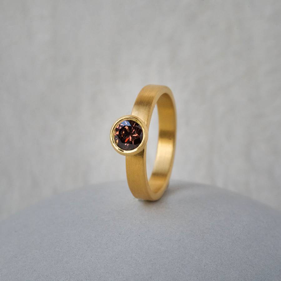 Minimalist yellow gold ring with a round cognac diamond in a bezel setting, displayed upright on a soft neutral surface with a light grey background.