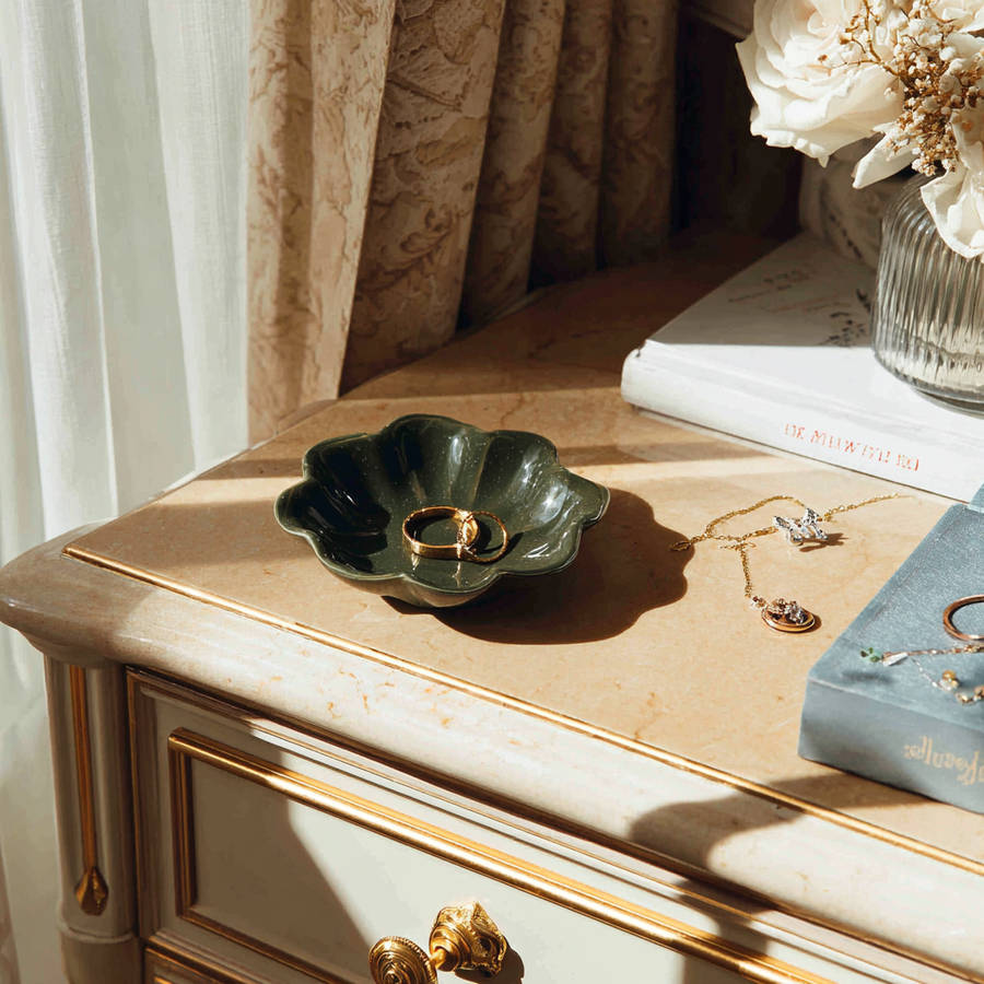 Two gold bespoke engagement rings in a green ceramic dish on a marble bedside table in sunlight.