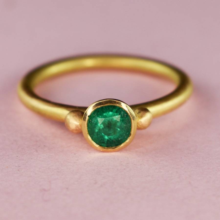 Gold bezel-set ring with a round green emerald centered between two small gold accents on a soft pink background