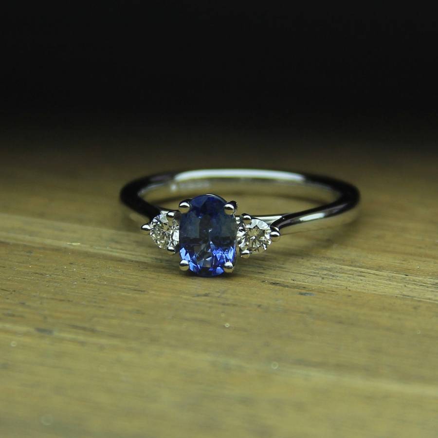 White gold engagement ring with an oval blue sapphire centre stone flanked by two round diamonds, resting on a wooden surface with a dark blurred background.
