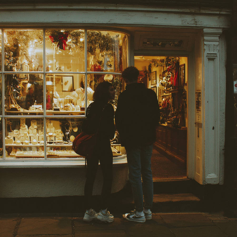 Couple window shopping at a warmly lit jewellery store at night, viewing rings and necklaces in festive display