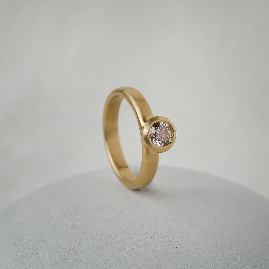 Bespoke yellow gold bezel-set engagement ring with a round champagne diamond, standing on a smooth pale stone against a soft neutral background