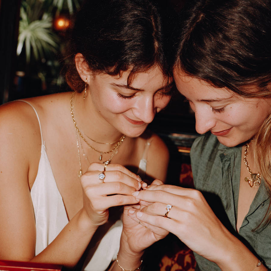 Two women smiling as they compare bespoke diamond engagement rings, holding their hands together in warm indoor light.