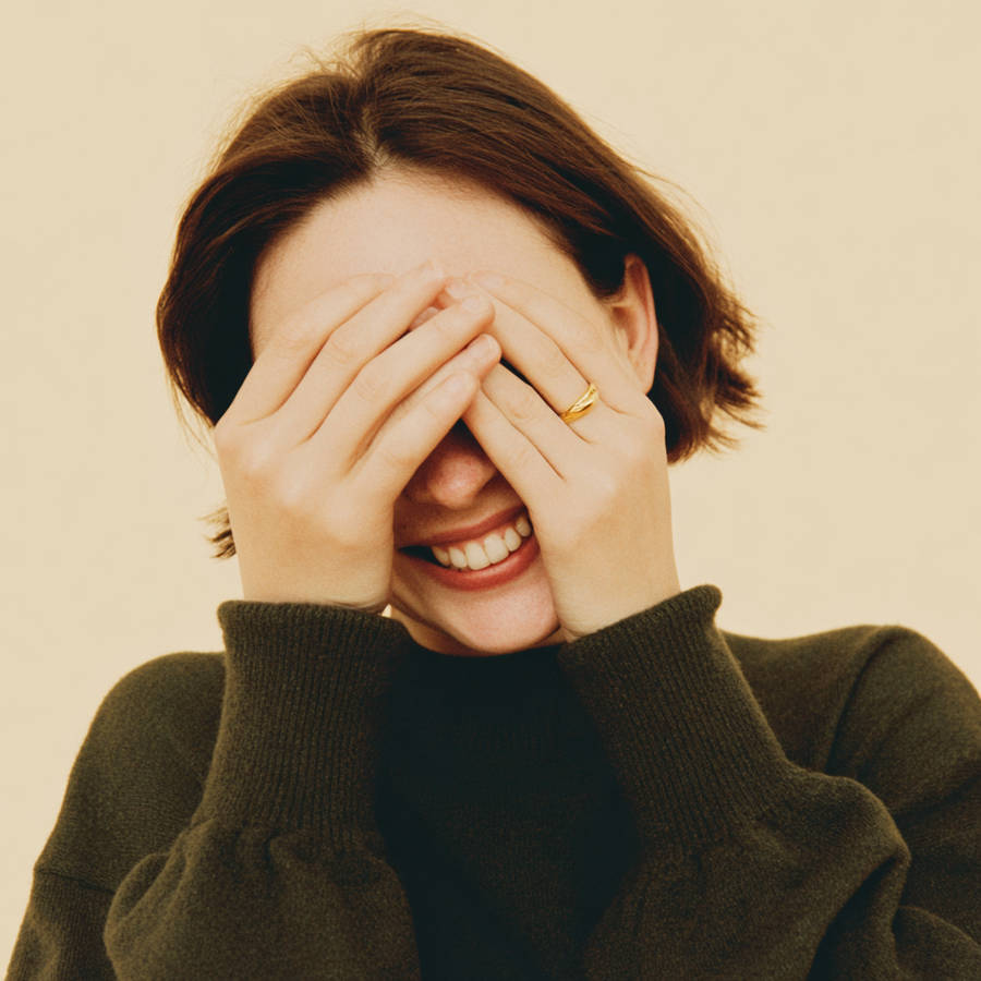 Smiling woman covering her face with hands, wearing a simple gold ring, warm soft lighting portrait