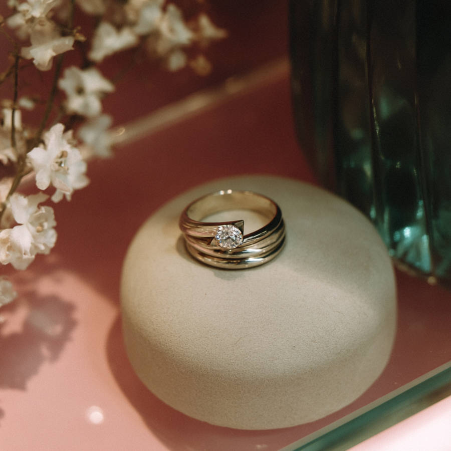 Bespoke white gold diamond ring resting on a stone display, with soft flowers and a glass bottle in the background.