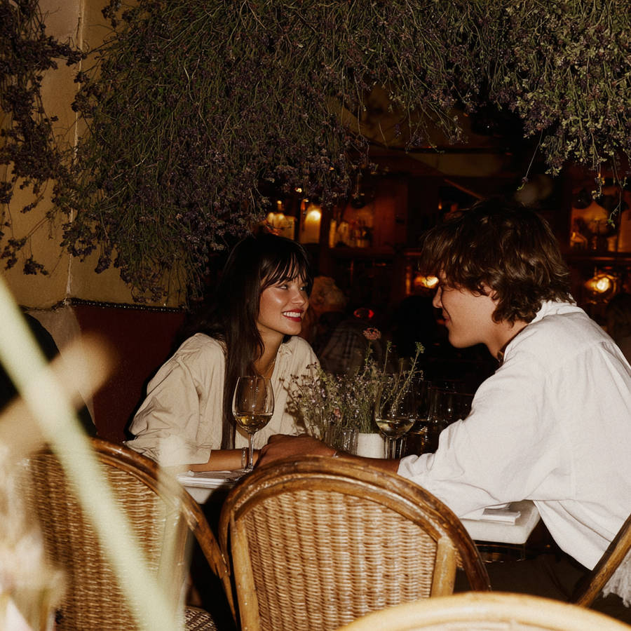 Couple smiling across a candlelit restaurant table with wine glasses, framed by hanging dried flowers overhead.