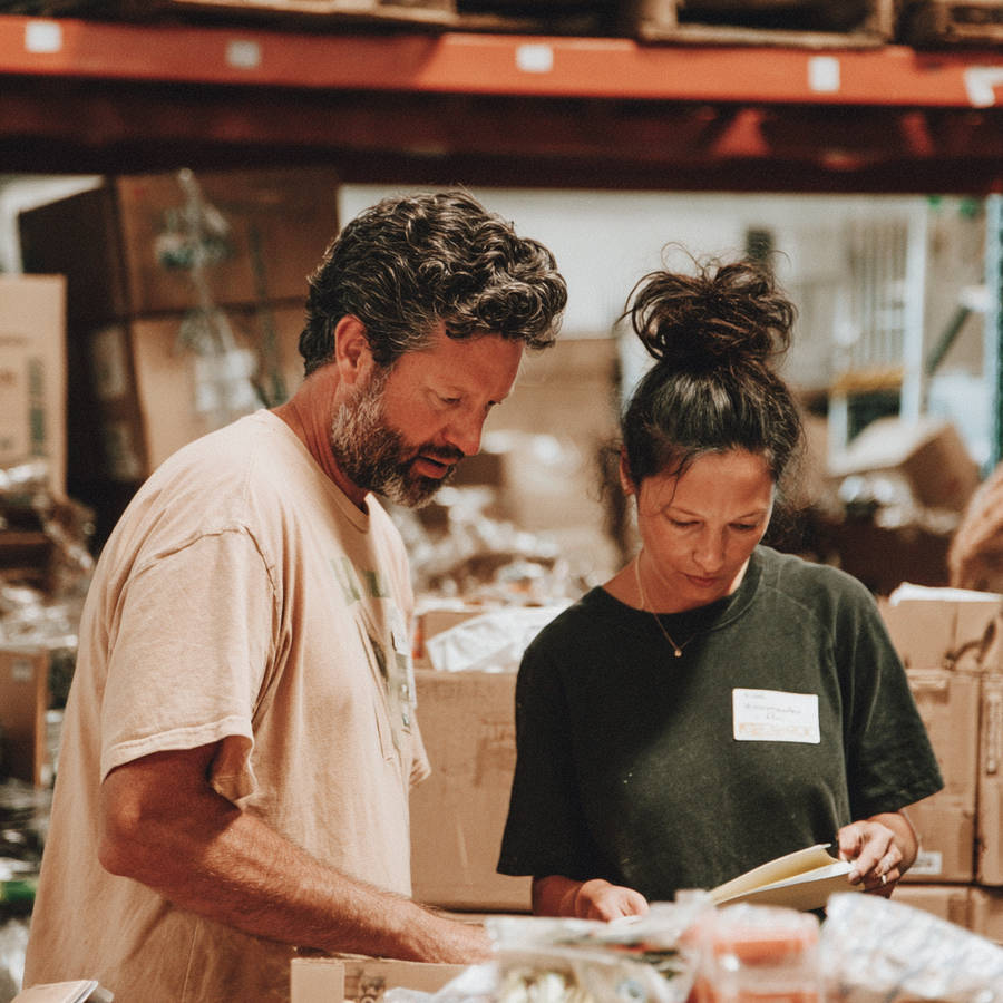 Man and woman volunteering at a food bank warehouse, sorting donations and checking a clipboard among stacked boxes.
