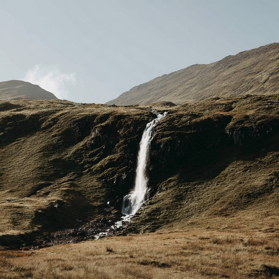 Slender waterfall cascading down mossy, grassy mountainside into a rocky stream beneath open sky in a remote natural landscape