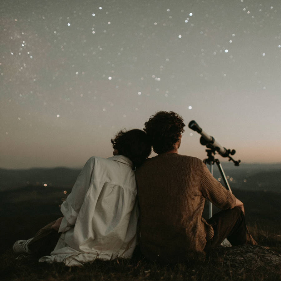 Couple sitting close while stargazing beneath a clear night sky, leaning together beside a telescope overlooking a quiet landscape