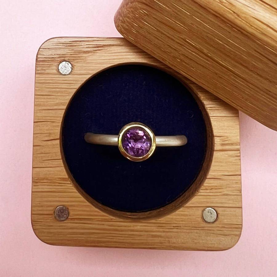 Gold ring with a round purple amethyst set in a bezel, displayed in an open wooden ring box on a pale pink background