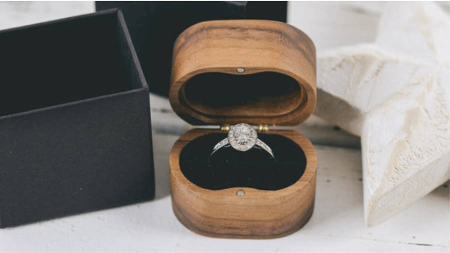 Diamond engagement ring in open wooden ring box beside black gift box on white linen background