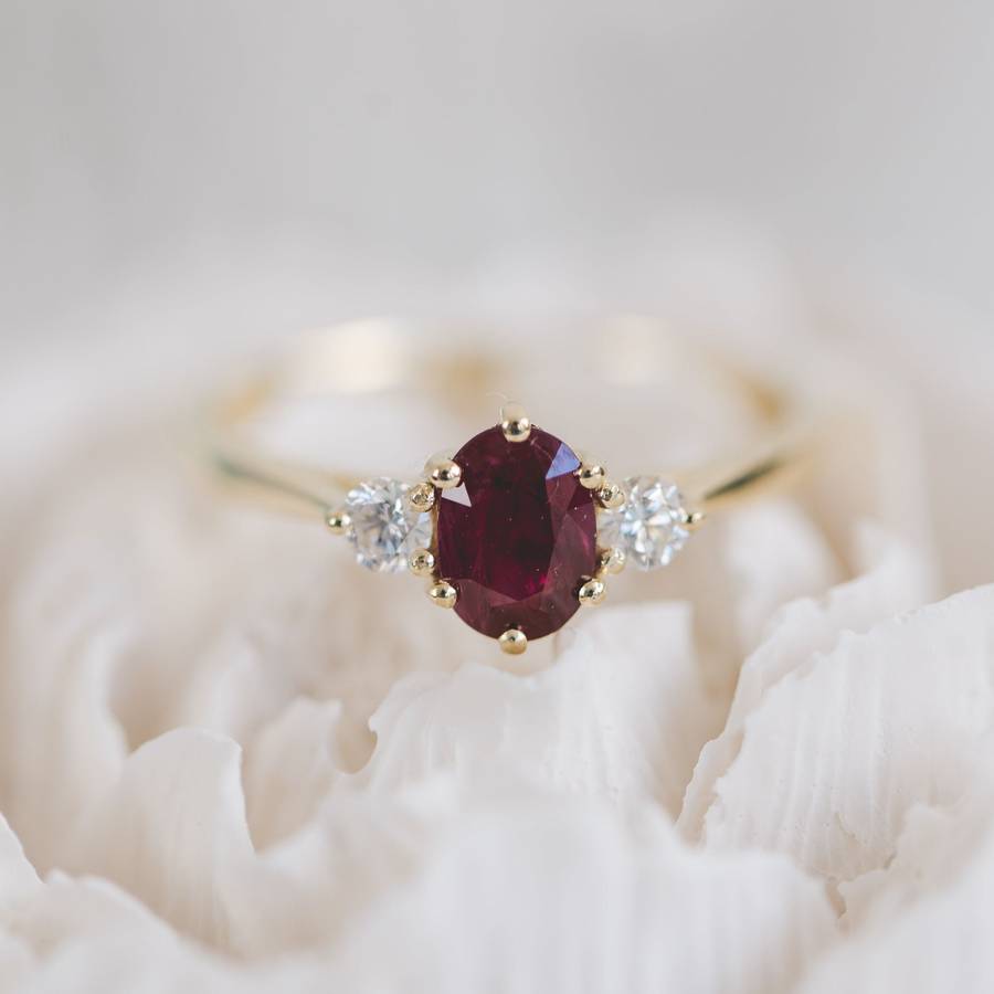 Yellow gold ring with an oval deep red ruby center stone flanked by two small round white diamonds, displayed among soft white seashell textures.