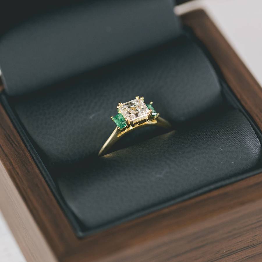 Bespoke yellow gold engagement ring with a square diamond and green side stones, presented in a dark leather ring box