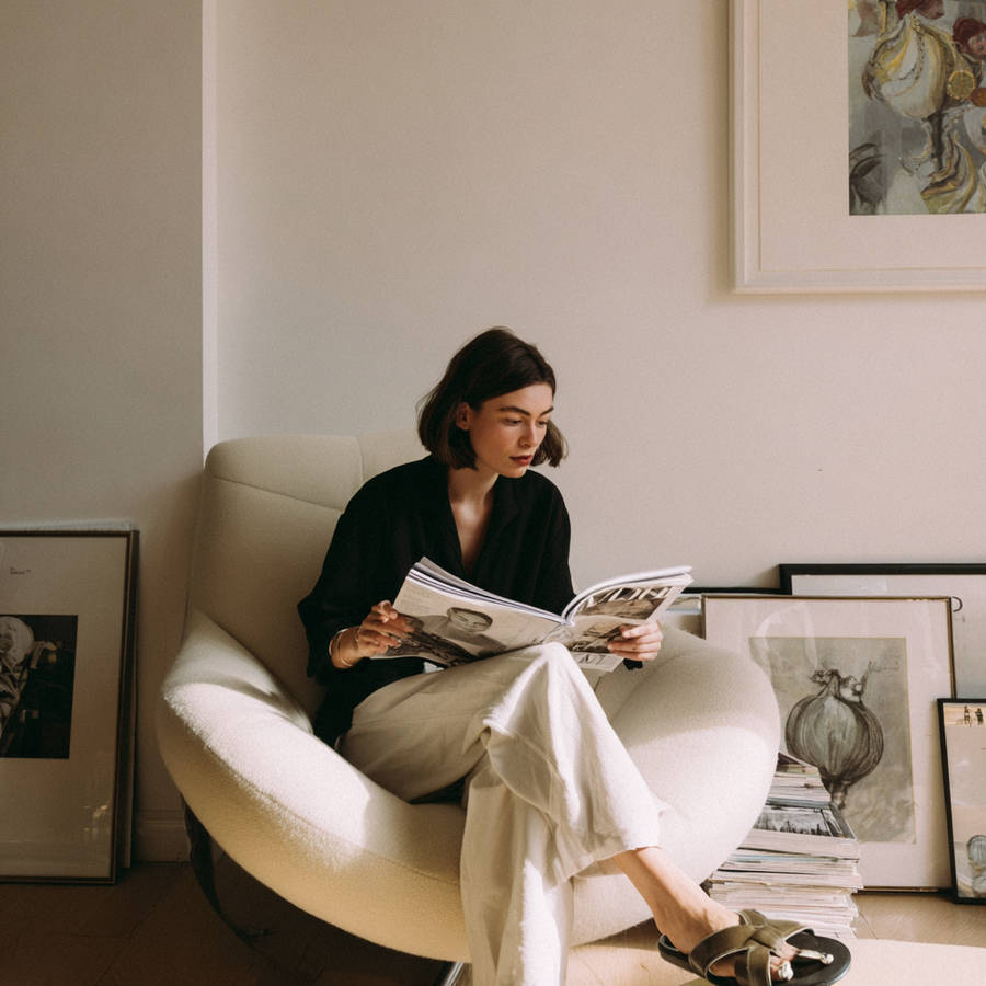 Woman in black top and white trousers reading a magazine in a cream armchair, framed artwork around her.