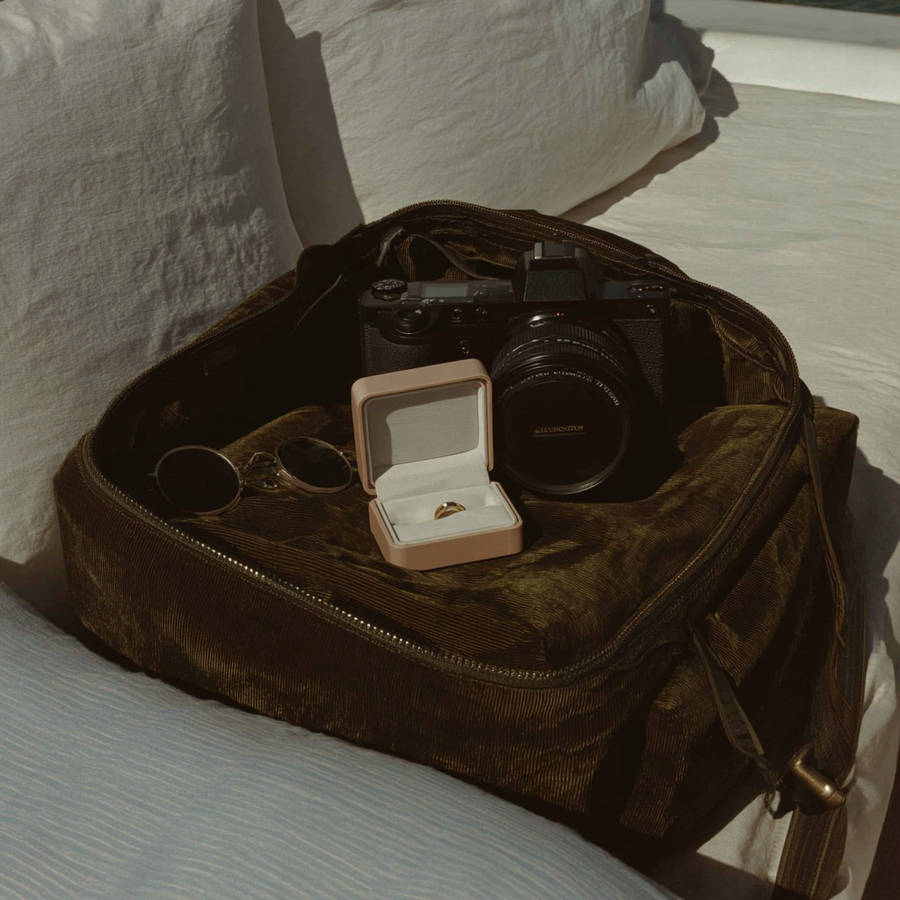 Open camera bag with photography gear, sunglasses and a ring box displaying a gold ring on a sunlit bed.