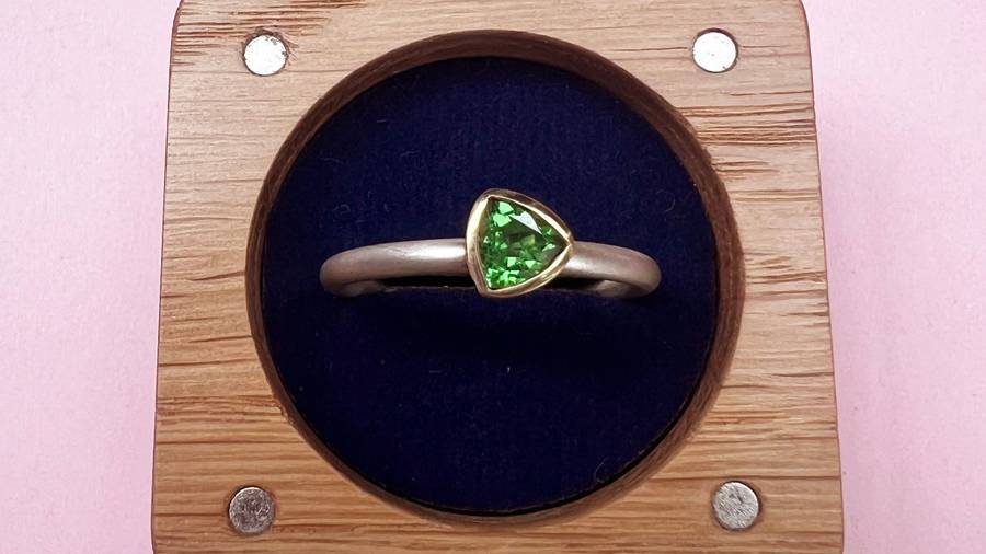 Minimalist silver ring with a small triangular green gemstone in a gold bezel setting, displayed in a wooden ring box with a dark blue interior against a pink background.