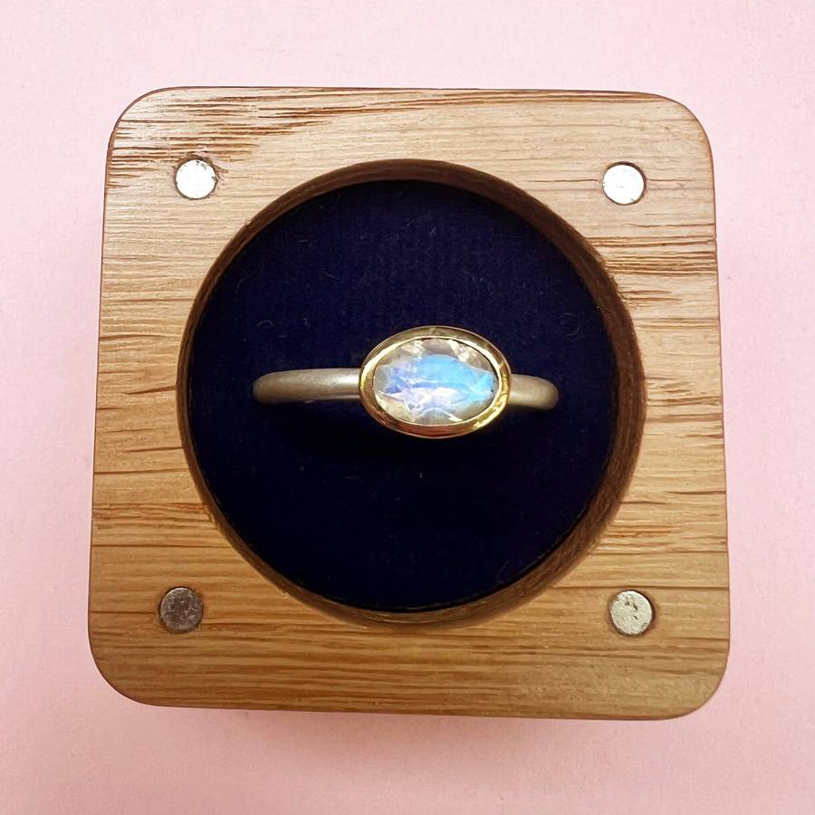 Bespoke yellow gold bezel-set ring with an oval moonstone, displayed in a wooden ring box with a dark velvet insert on a soft pink background
